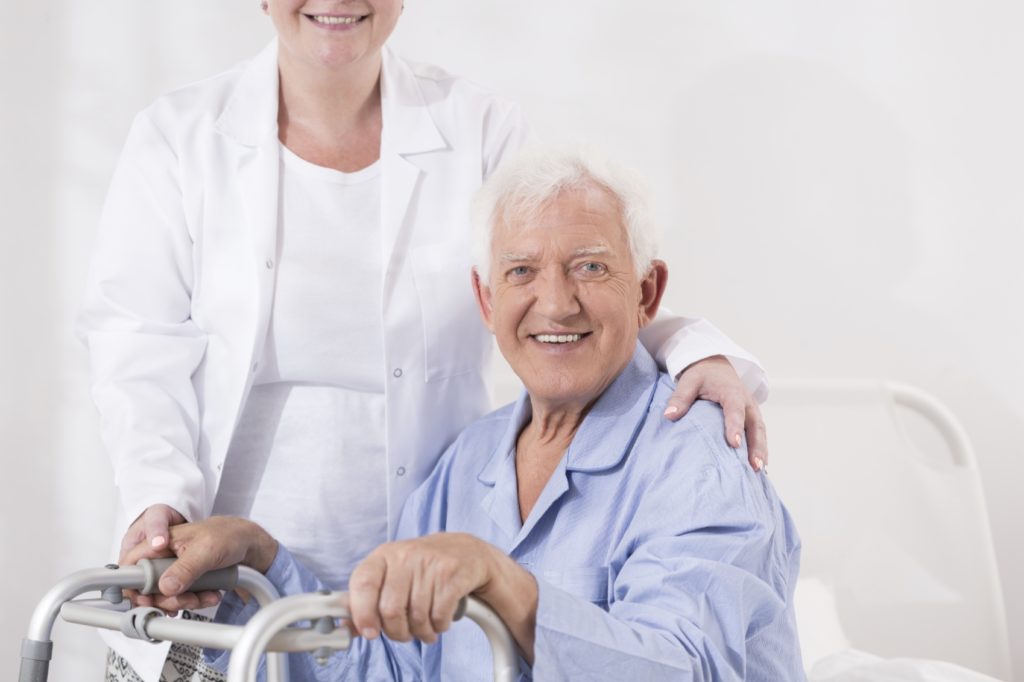 An older adult sits down with his hands on a walker while a woman providing ALS care stands behind him with her hand on his shoulder.