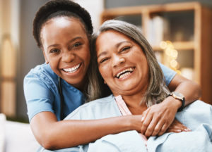 A smiling female caregiver who provides 24-hour in-home care hugs her happy client.