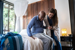 A female caregiver providing live-in care for older adults assists an older man in his home.